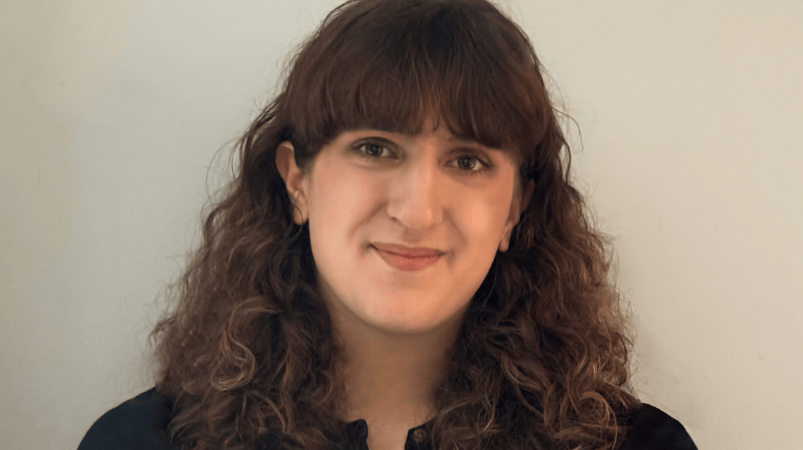 Young woman smiling with curly brown hair and bangs, wearing a dark top, in front of a light-colored background.